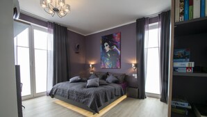 A modern bedroom with a large bed, a painting on the wall, and a bookshelf with books and a box.