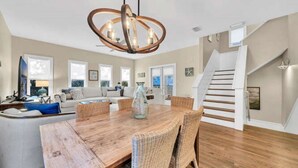 A spacious living room with a wooden dining table, wicker chairs, a large chandelier, and a staircase leading to an upper level. - Above the Sea in Old Florida Cottages. South of 30a, Sunset views, Mins to beach (Santa Rosa Beach)