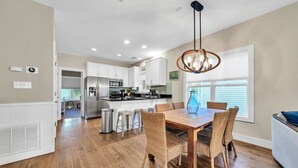 A modern kitchen with white cabinets, stainless steel appliances, and a dining area with a wooden table and wicker chairs. - Above the Sea in Old Florida Cottages. South of 30a, Sunset views, Mins to beach (Santa Rosa Beach)