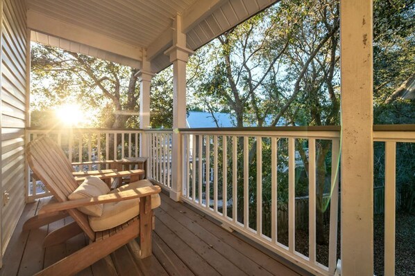 A wooden porch with a white railing, a wooden rocking chair, and a view of trees and a house in the background.
