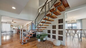 A spacious living area with a wooden staircase, a dining area with white chairs, and a kitchen in the background. - Horseshoe Bay - Lakefront + Pool - Lucy on the Lake (Horseshoe Bay)