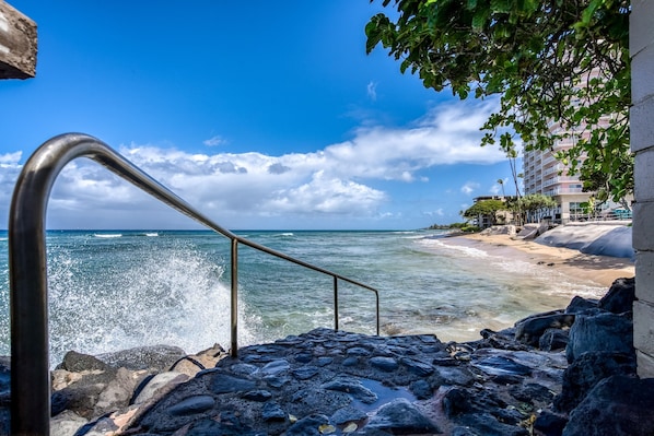 A staircase leading to a beach with waves crashing onto the shore, a clear sky, and a building with balconies in the background.