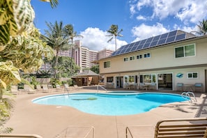 A swimming pool with lounge chairs, a covered patio, and a building with solar panels.