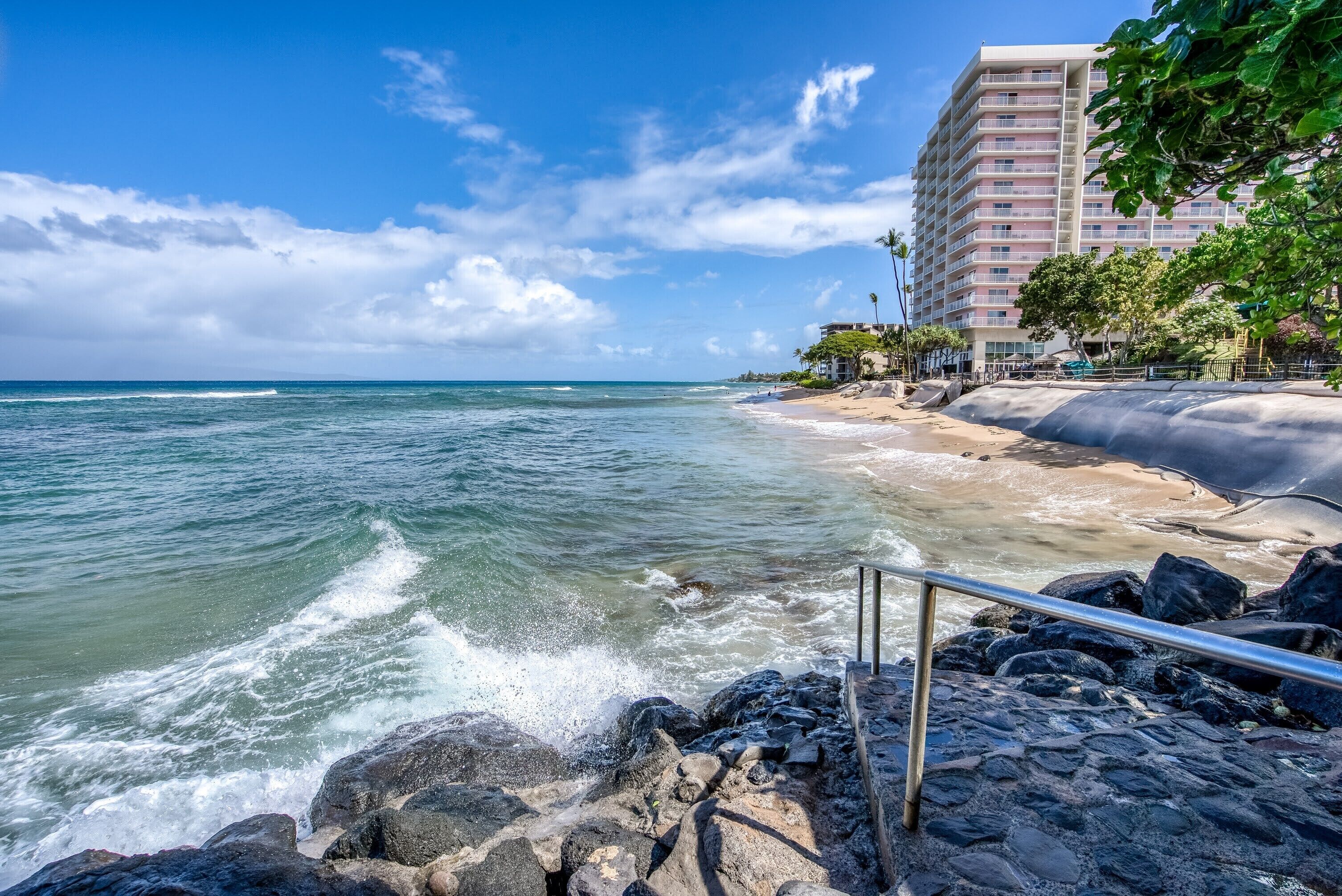A beach with waves crashing onto rocks, a metal railing, and a multi-story building with balconies in the background.