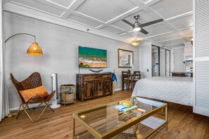 Ein modernes Schlafzimmer mit einem Bett, einem Glastisch, einer Holzkommode, einem Sessel, einem Deckenventilator und einem Bild an der Wand.