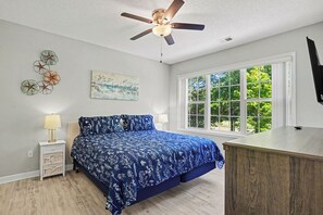 A bedroom with a bed, a ceiling fan, a window with a view, a nightstand, and a dresser.