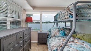 Een stapelbed met een gebedrukte spreideken, een dressoir met lades, een nachtkastje met een lamp en een raam met jaloezieën met uitzicht op een wateroppervlak.
