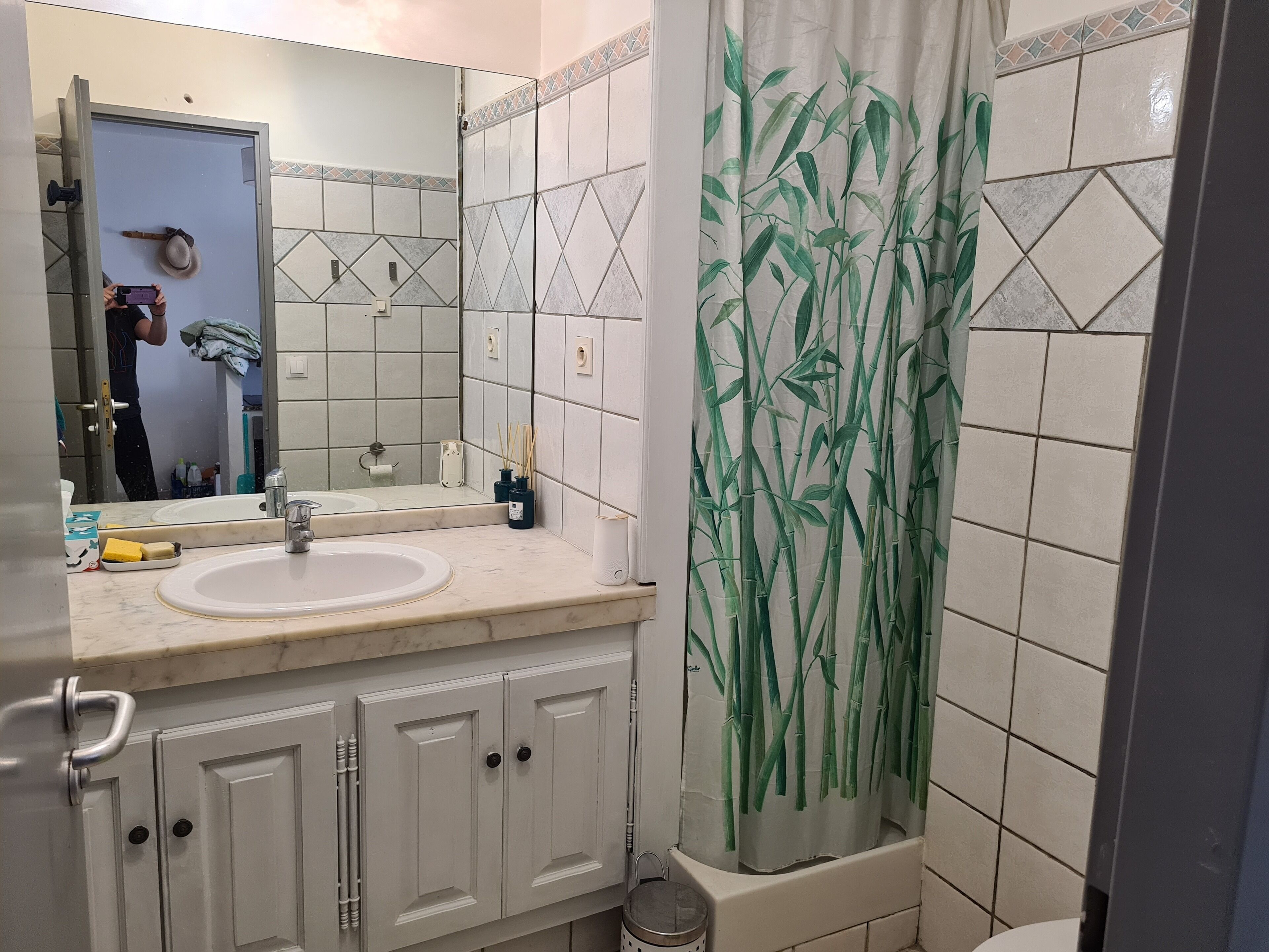 Une salle de bain avec un lavabo blanc, un miroir et des armoires. Il y a un rideau de douche orné d’un motif en bambou.