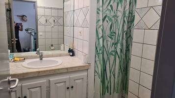 Une salle de bain avec un lavabo blanc, un miroir et des armoires. Il y a un rideau de douche orné d’un motif en bambou.