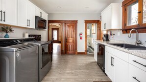 A modern kitchen with white cabinets, stainless steel appliances, and a wooden door. - Beautiful House | Walk Everywhere | AC| Fire Pit | (Traverse City)
