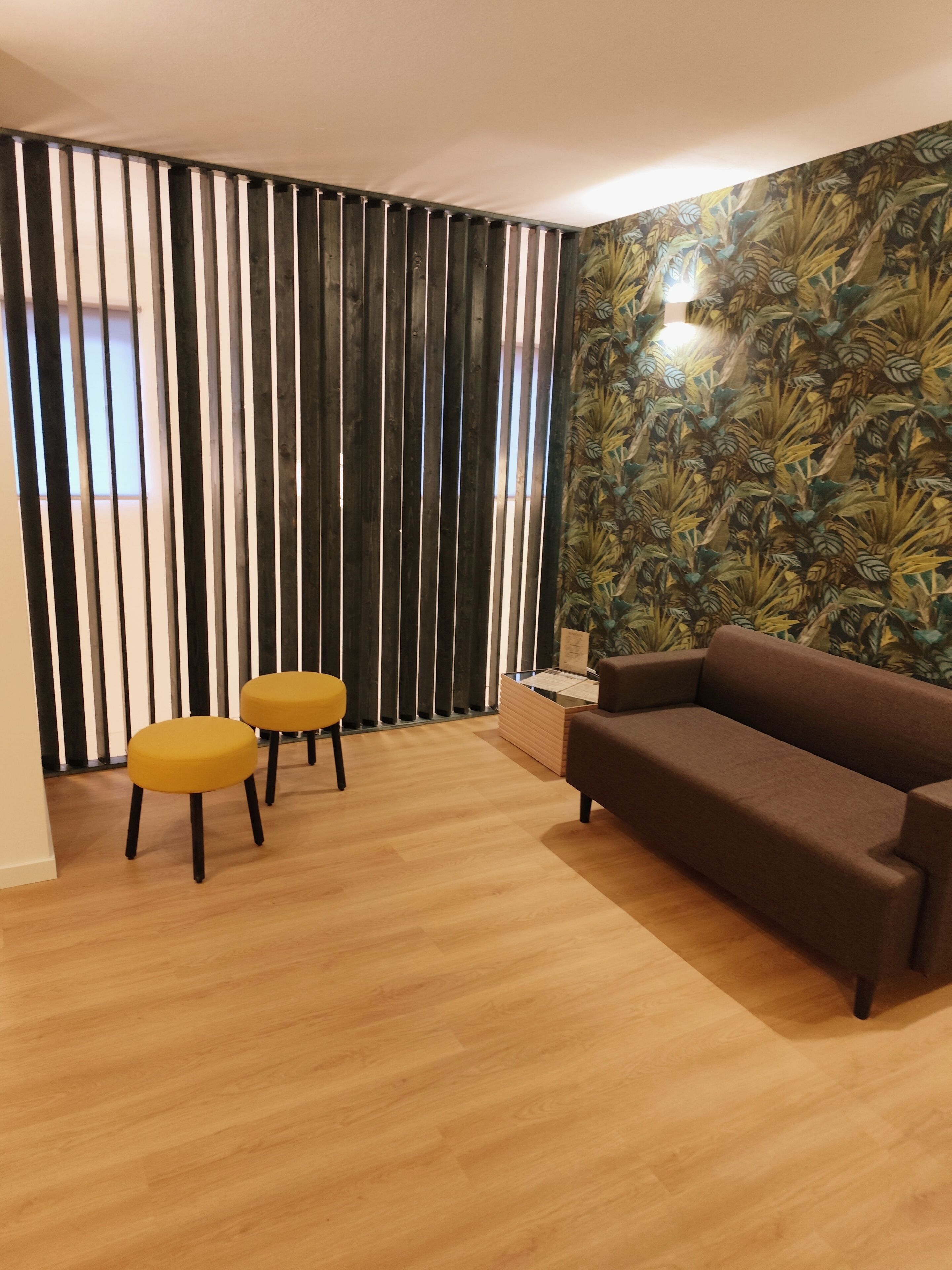 Uma sala de estar com um sofá marrom, dois banquinhos amarelos, piso de madeira, um divisória de venezianas verticais e um papel de parede tropical.