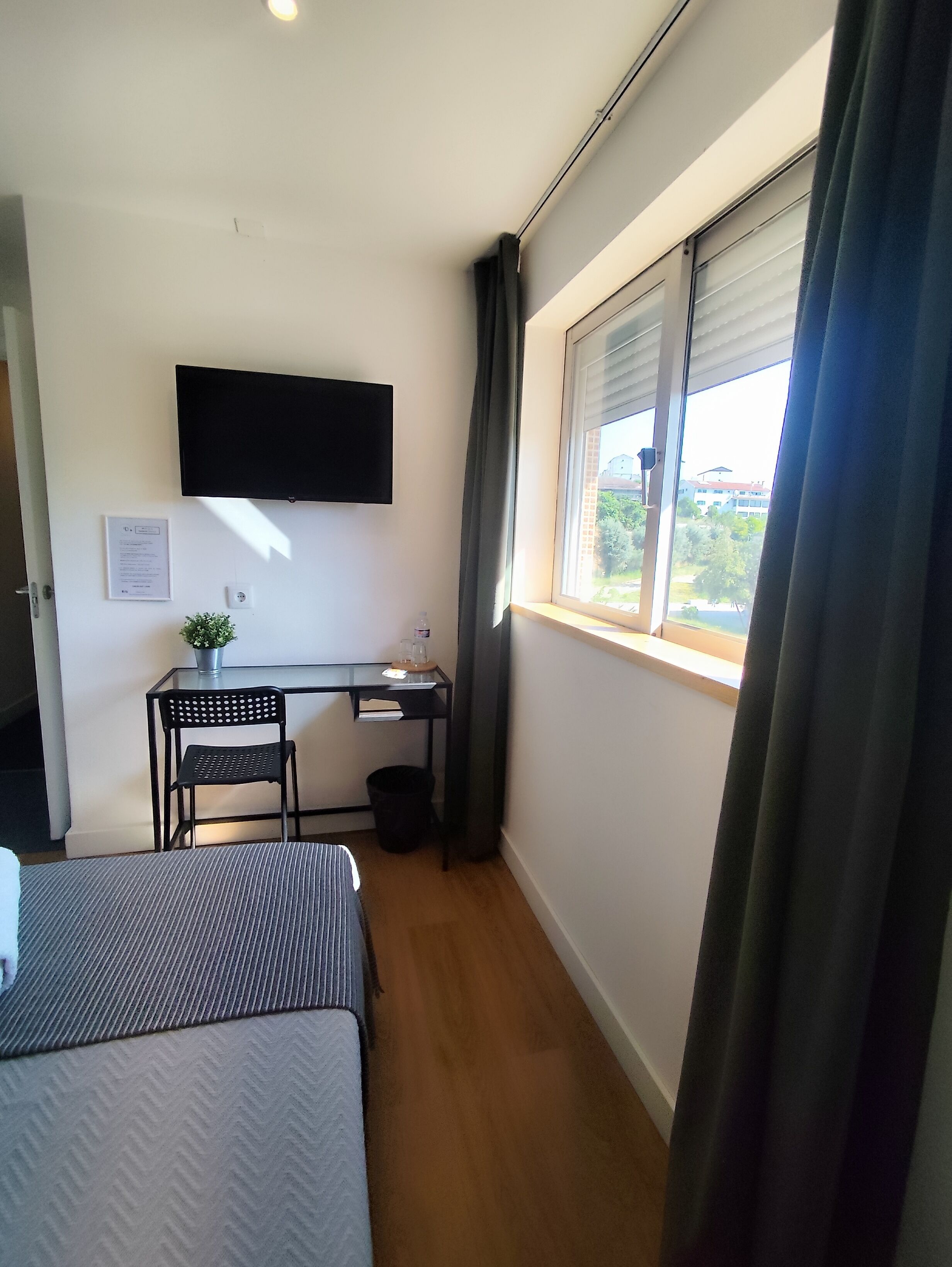 Una habitación pequeña con una cama, un escritorio, una silla, un televisor montado en la pared y una ventana con cortinas.