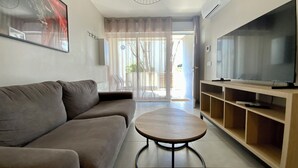 A living room with a sofa, a round coffee table, a TV on a wooden stand, and a sliding glass door leading to an outdoor area.