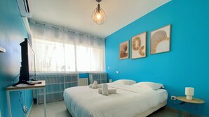 A bedroom with a bed, a desk with a computer, a window with sheer curtains, and wall art.