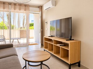 A living room with a flat-screen TV on a wooden stand, a sofa, a round table, and a sliding glass door leading to an outdoor area.