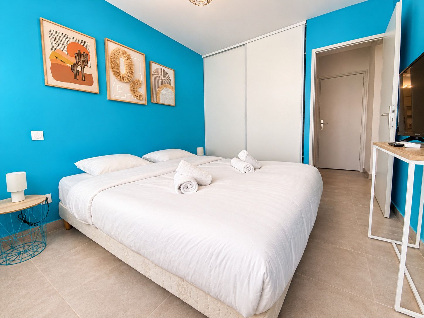 A hotel room with a large bed, two side tables, a desk, and artwork on the wall.