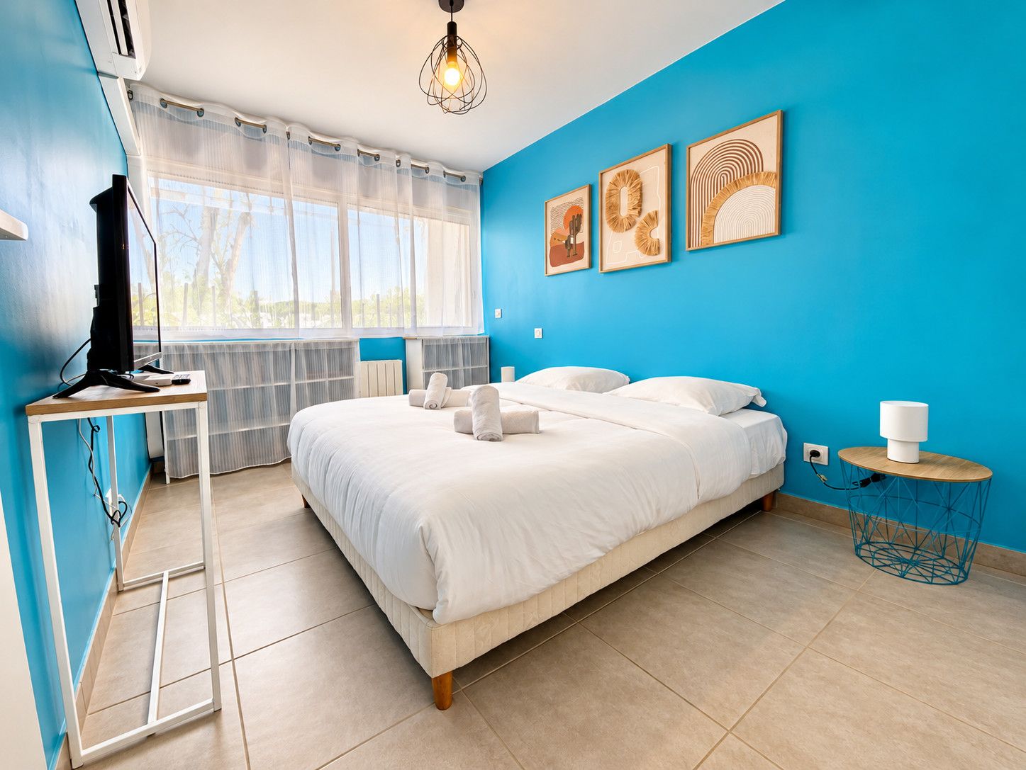 A bedroom with a bed, a desk with a computer, a bedside table, and artwork on the wall.
