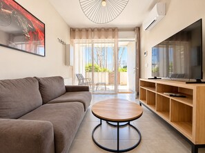 A modern living room with a sofa, a round coffee table, a TV on a wooden stand, and a sliding glass door leading to an outdoor area.