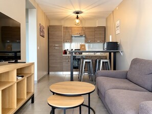 A modern living room with a sofa, two round tables, a TV, and a kitchen area with wooden cabinets and a hanging light fixture.
