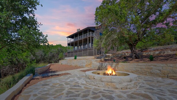 A stone patio with a fire pit, surrounded by a stone wall and greenery.