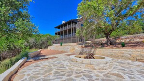 A stone patio with a fire pit, surrounded by a stone wall and a wooden deck with a balcony overlooking a landscaped area with trees and shrubs.