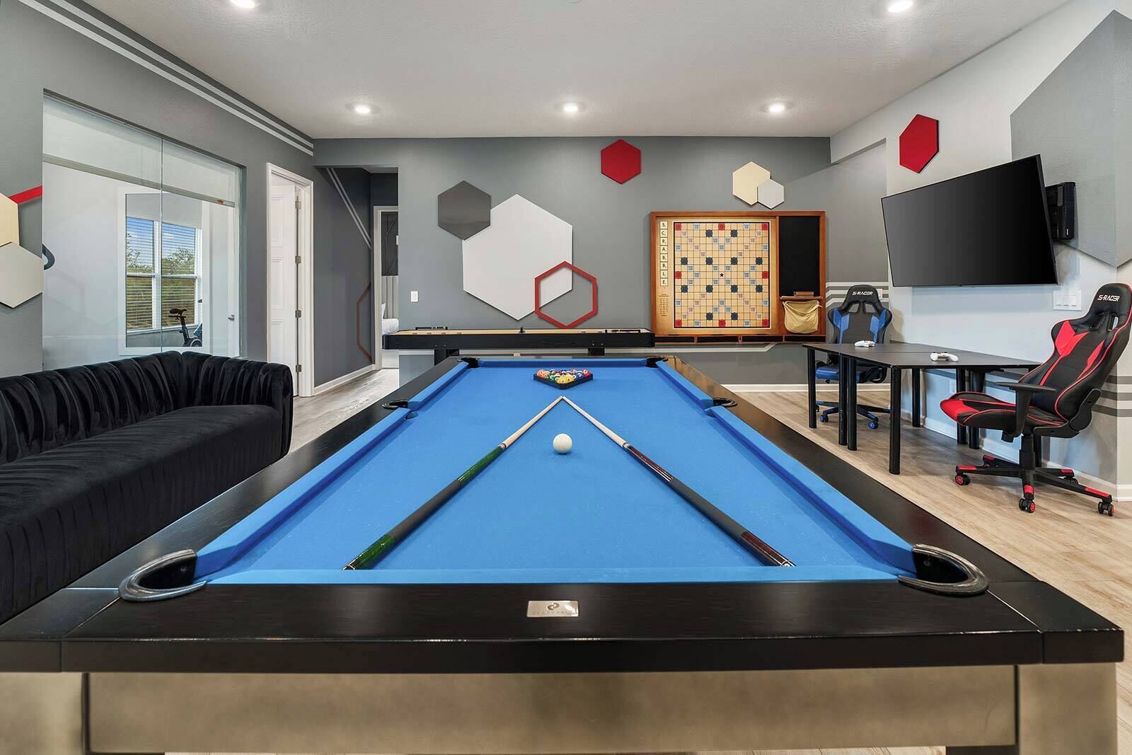 Een pooltafel met een blauwe viltkleed, een zwarte bank, een speeltafel met stoelen en een wandtelevisie.