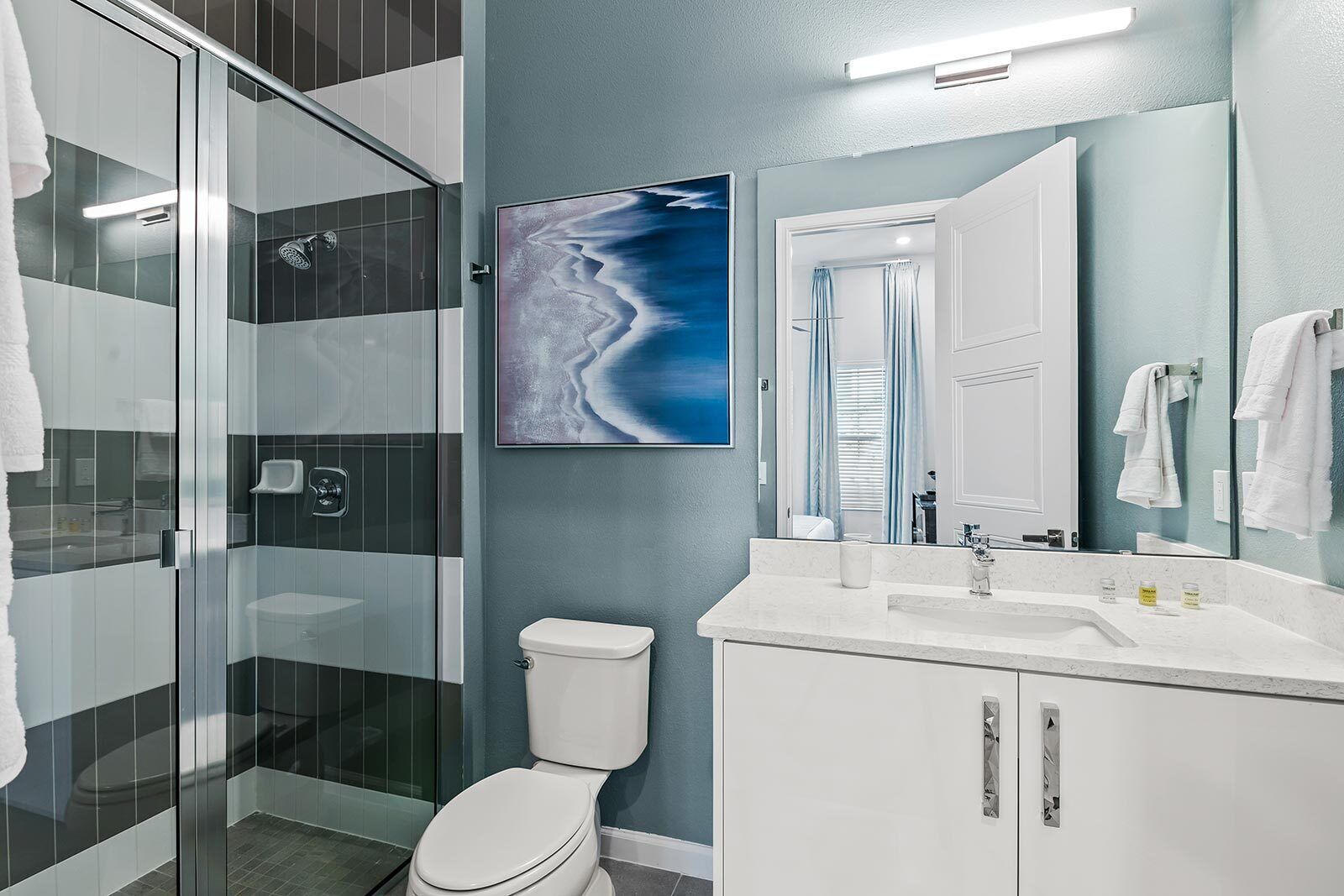 Un baño moderno con cabina de ducha de vidrio, inodoro blanco, lavamanos con espejo y un cuadro abstracto azul en la pared.