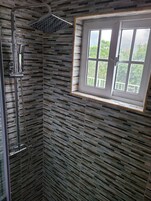 A shower with a rain showerhead and handheld showerhead, a window with a view of greenery, and a patterned wall.
