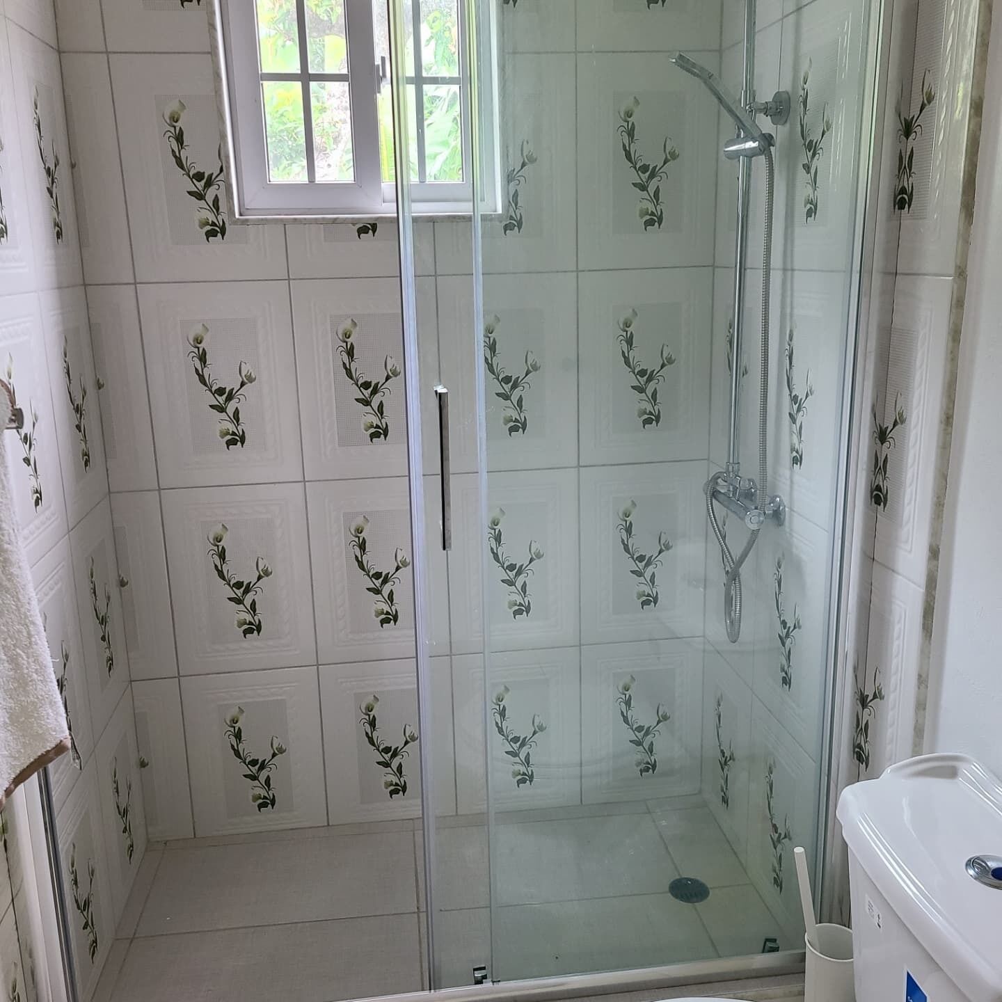 A bathroom with a glass shower enclosure, white tiled walls with floral patterns, a window above the shower, and a white sink with a blue label.