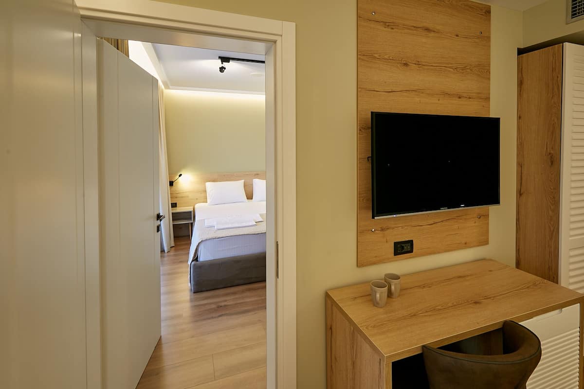 A hotel room with a wooden floor, a bed with white linens, a mounted TV, and a small wooden console table with a cup.