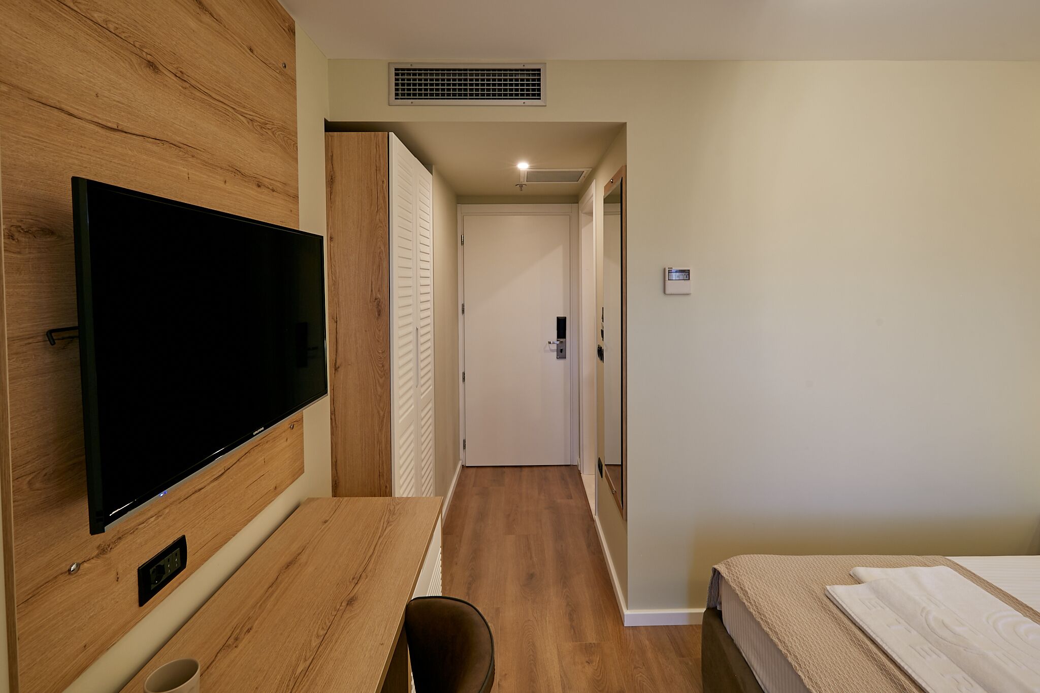 A hotel room with a wooden wall-mounted TV, a bed with a white mattress, and a wooden floor.