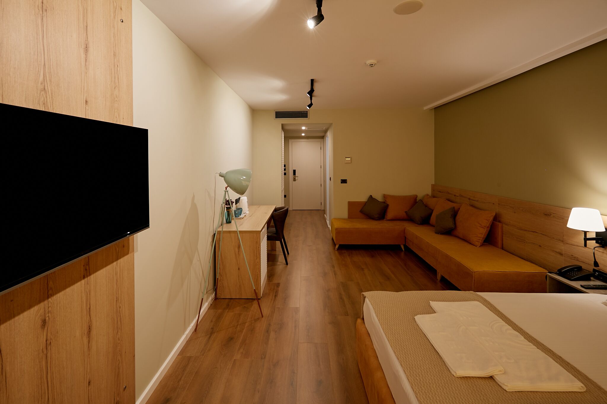 Una habitación de hotel con suelo de madera, una cama, un escritorio con lámpara, un sofá y un televisor en la pared.