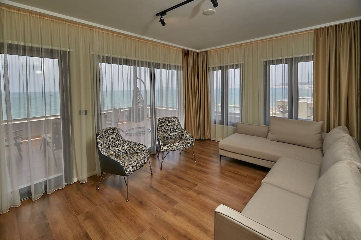 A spacious living room with a large sofa and two patterned chairs, a balcony with a glass door, and a view of the sea.