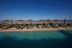Zona de piscina con tumbonas y sombrillas, cielo azul despejado y vista al mar.