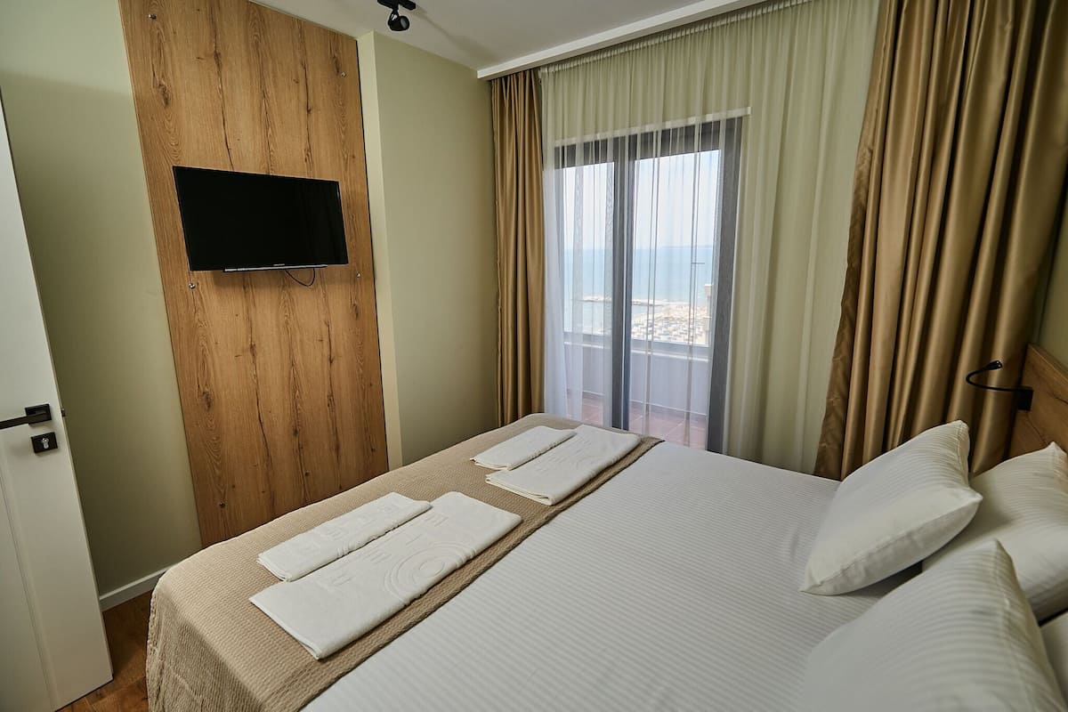 A hotel room with a bed, a TV mounted on a wooden wall, and a balcony with curtains.