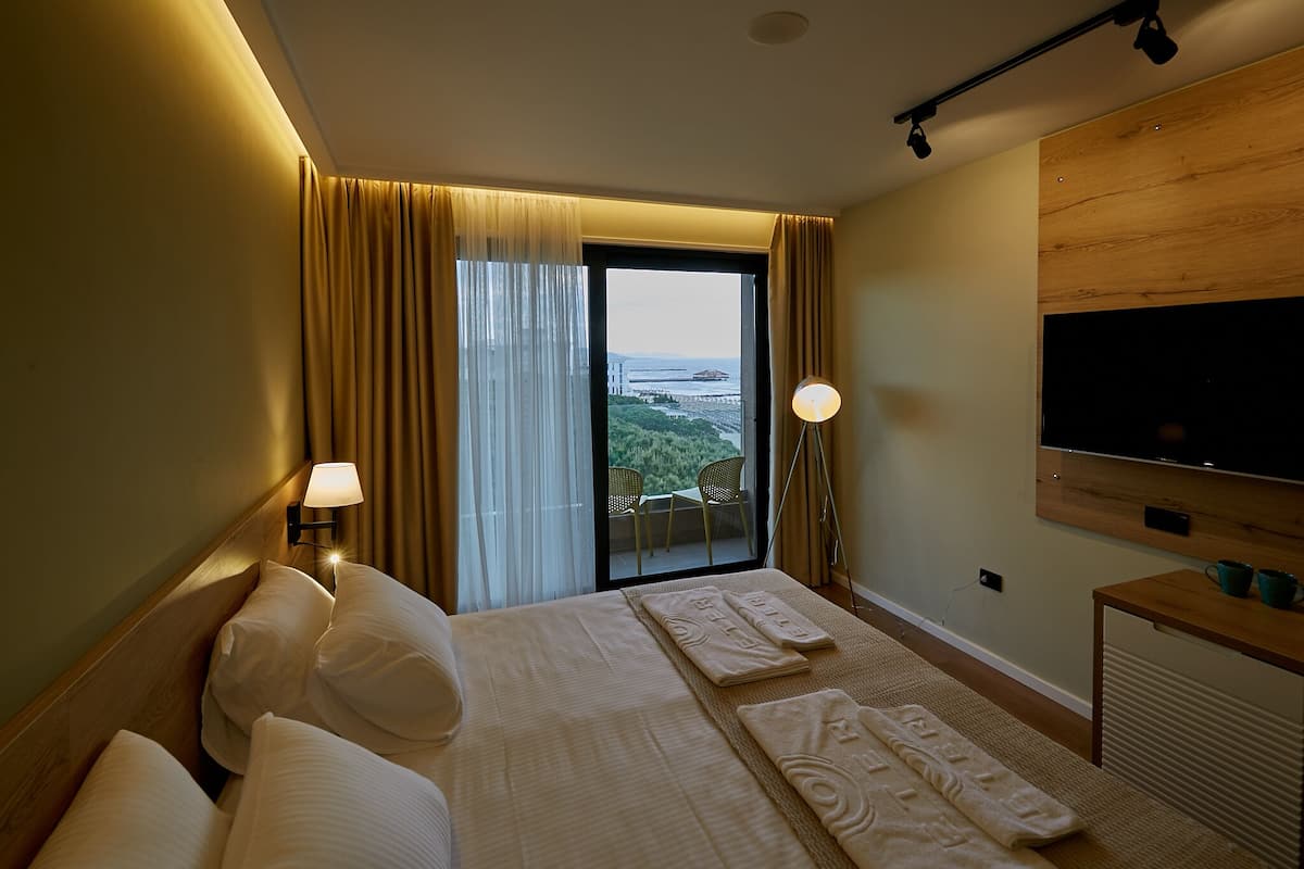 A hotel room with a large bed, two bedside lamps, a flat-screen TV, and a view of the outdoors through a large window.