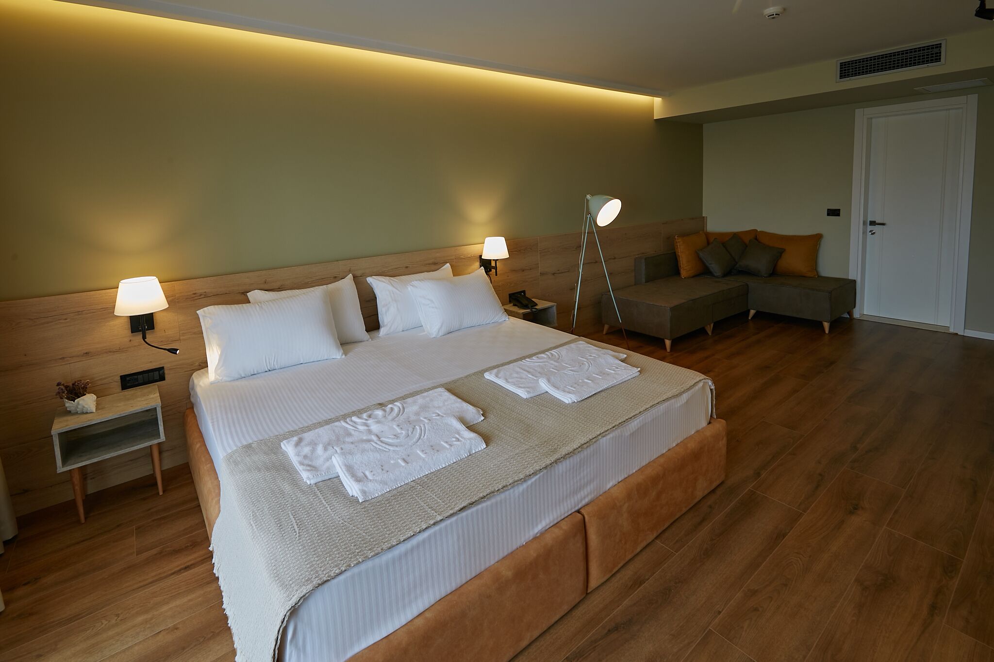 A hotel room with a large bed, two bedside tables, a nightstand, a sofa, and a door.