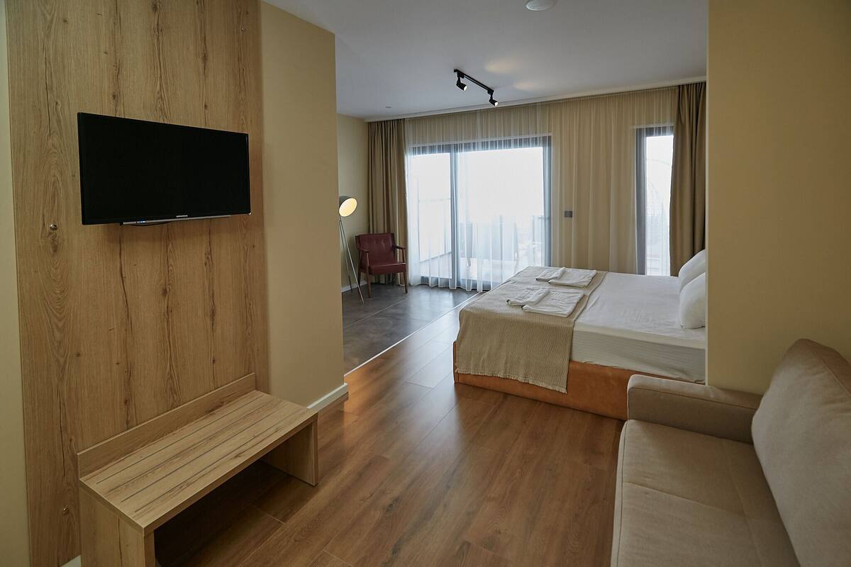 A hotel room with a bed, a TV mounted on a wooden wall, a bench, and a view of the city through large windows.