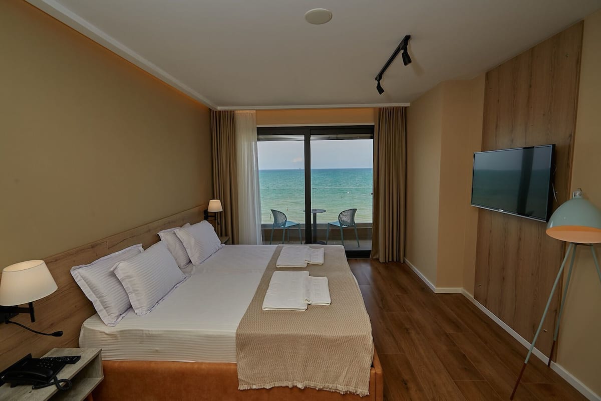 A hotel room with a bed, a TV, a desk with a phone, and a view of the sea through a large window.