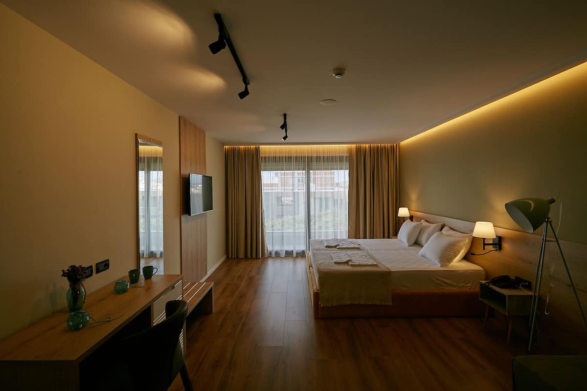 A hotel room with a bed, bedside table, lamp, TV, and a view of the outdoors through large windows.