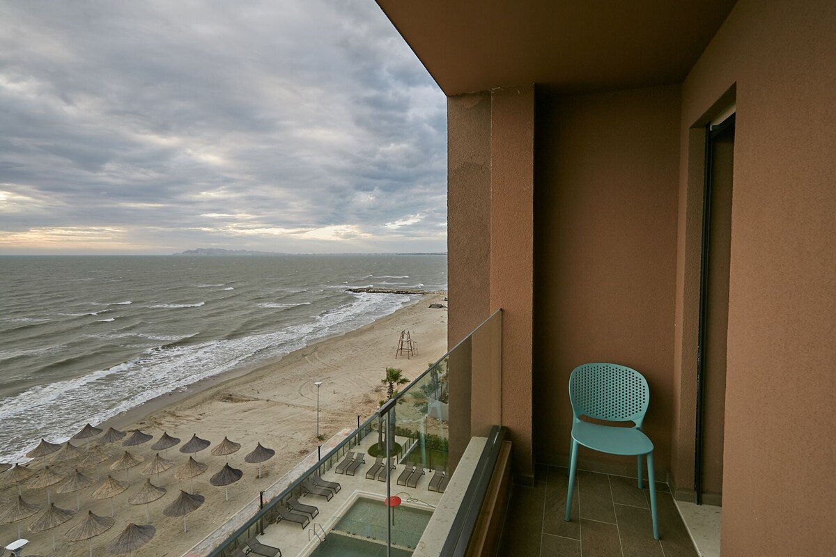 A balcony with a view of the beach and ocean, a chair, and a pool area.