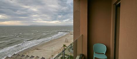 A balcony with a view of the beach, a chair, and a pool area.