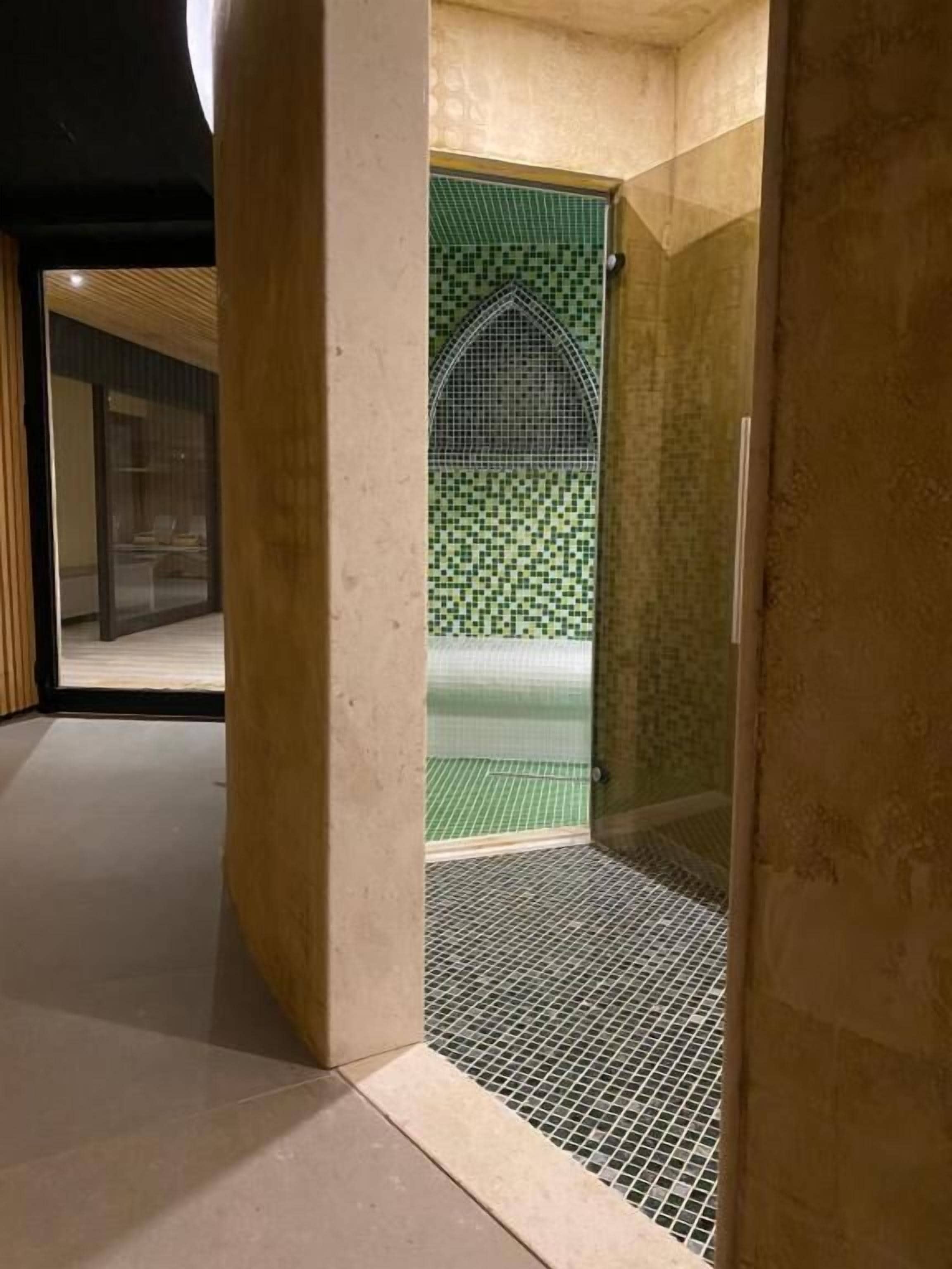 A modern bathroom with a green mosaic wall, a shower area with a metal grate, and a view of a wooden deck outside.