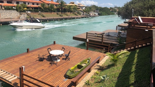 A wooden deck with a table and chairs, a green boat, and a waterway with a boat in the distance.