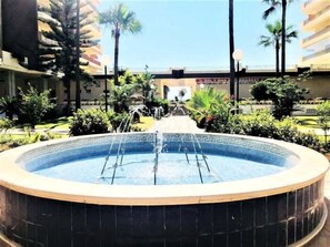 A circular pool with a central water feature, surrounded by greenery and palm trees, with a building in the background.