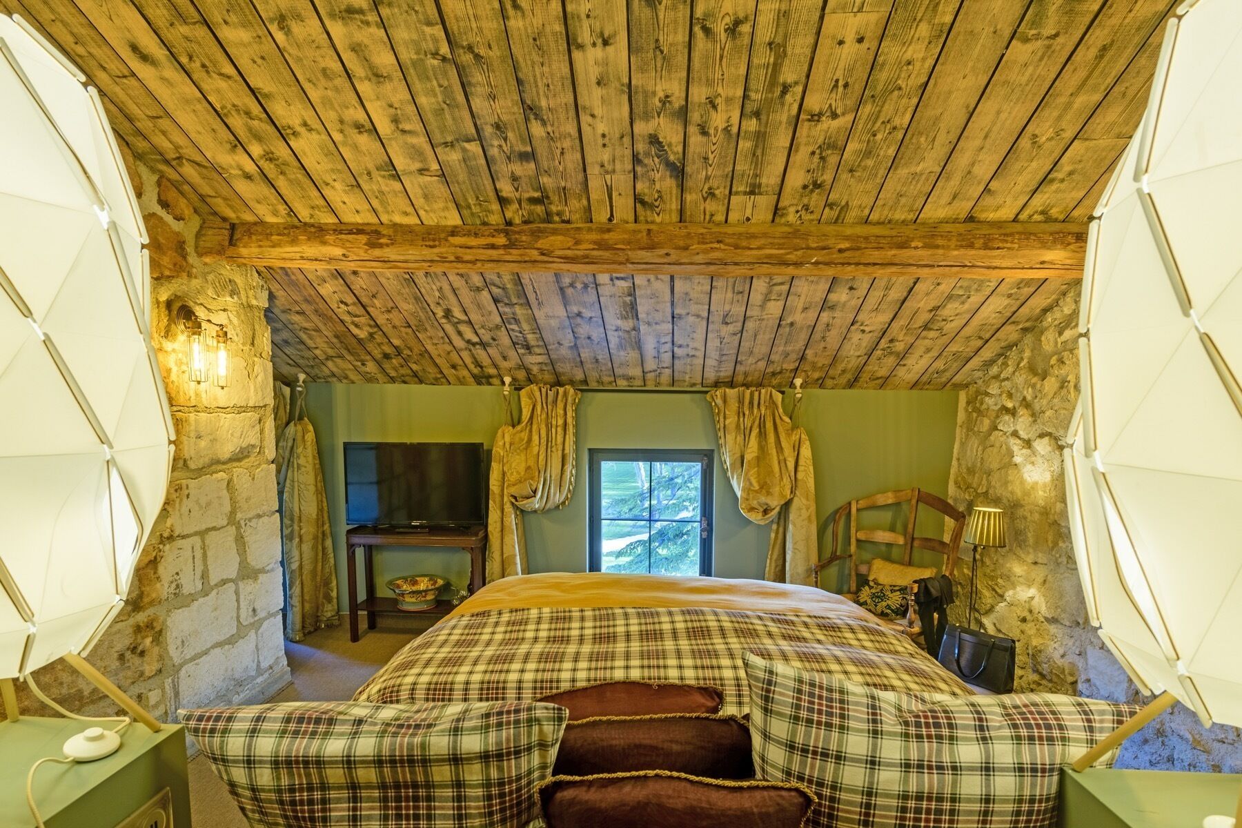 Um quarto rústico com teto de madeira, cama com roupa de cama xadrez, parede de pedra e janela com cortinas.