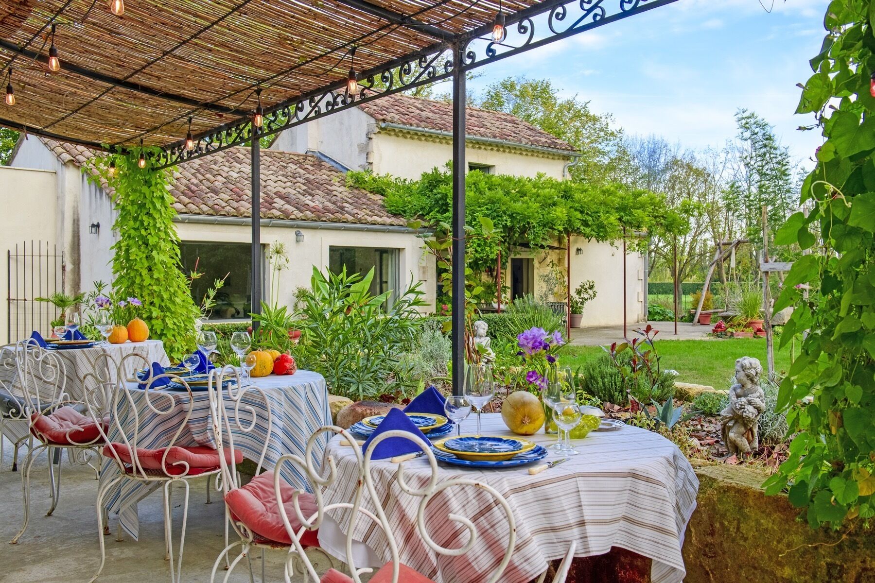 An outdoor dining area with tables set for a meal, surrounded by greenery and a house in the background.
