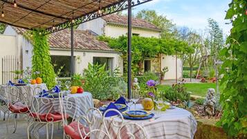 An outdoor dining area with tables set for a meal, surrounded by greenery and a house in the background.