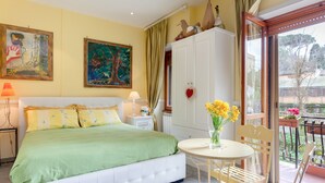 Una camera da letto con un letto, una comodino, tende a una finestra, un balcone con tavolo e sedie, e un quadro appeso al muro.