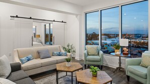 A modern living room with a large window, a sofa, two armchairs, a coffee table, and a side table with a lamp. - Boardwalk Pent House | Ocean Views + Beach Access! (Carolina Beach)